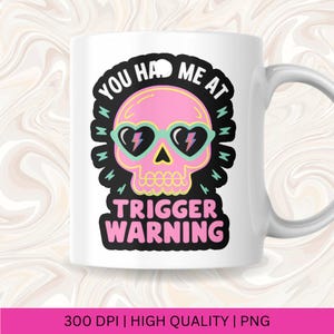 Pu&ograve; includere: Tazza in ceramica bianca con un disegno di teschio rosa che indossa occhiali da sole a forma di cuore con dettagli a fulmine. La tazza ha il testo "YOU HAD ME AT TRIGGER WARNING" in nero e rosa. La tazza ha un manico bianco.