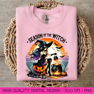 May include: A pink sweatshirt featuring a graphic design of a witch stirring a cauldron. The text "Season of the Witch" is written in a black and white font above the witch. The witch is wearing a black dress and a black hat with a purple bow. The background is a sunset with orange, pink, and purple colours.