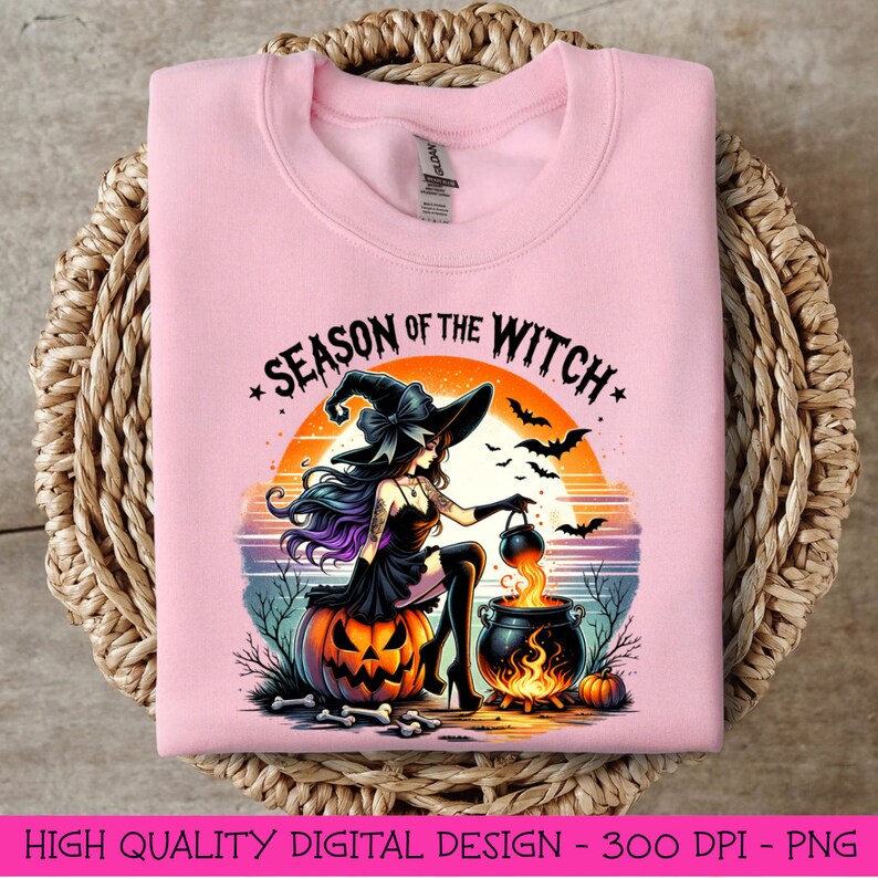 May include: A pink sweatshirt featuring a graphic design of a witch stirring a cauldron. The text "Season of the Witch" is written in a black and white font above the witch. The witch is wearing a black dress and a black hat with a purple bow. The background is a sunset with orange, pink, and purple colours.