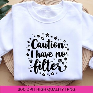 Puede incluir: Sudadera blanca con texto negro que dice "Cuidado, no tengo filtro" rodeado de peque&ntilde;as flores negras.