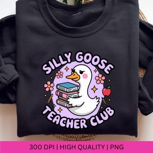 Op de afbeelding: Zwart sweatshirt met een afbeelding van een witte gans die boeken vasthoudt, omringd door bloemen, sterren en een appel. De tekst luidt "Silly Goose Teacher Club".