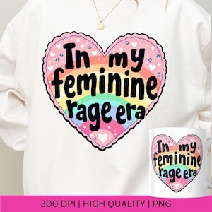 May include: A white sweatshirt with a rainbow heart design. The heart has the text "In my feminine rage era" in black lettering.