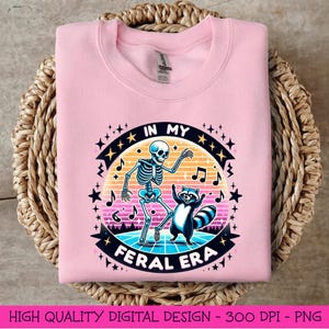 May include: Pink sweatshirt featuring a graphic design of a skeleton and a raccoon dancing with the text "In My Feral Era" in a retro style.