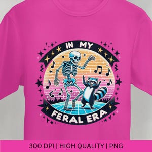 May include: A pink t-shirt featuring a graphic design of a skeleton and a raccoon dancing on a retro-style dance floor. The text "In My Feral Era" is written in a circle around the dancing figures.