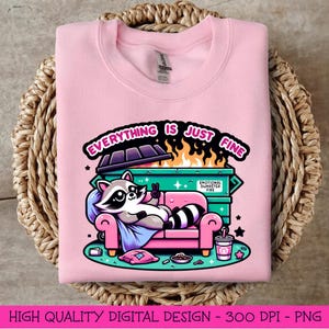 May include: A pink sweatshirt with a graphic design of a raccoon sitting on a couch in front of a dumpster fire. The text "Everything is just fine" is written above the dumpster.