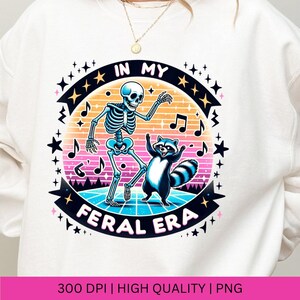 May include: White sweatshirt featuring a graphic of a skeleton and a raccoon dancing with the text "In My Feral Era" in a retro style.