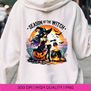 May include: A white hoodie featuring a graphic print of a witch stirring a cauldron against a sunset backdrop. The text "Season of the Witch" is written in black and white with stars on either side.
