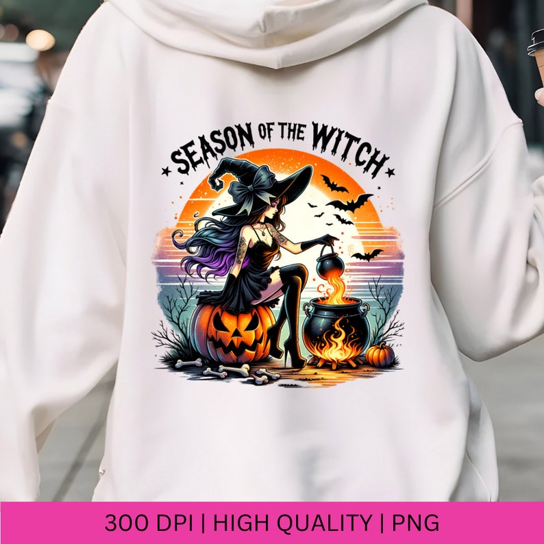 May include: A white hoodie featuring a graphic print of a witch stirring a cauldron against a sunset backdrop. The text "Season of the Witch" is written in black and white with stars on either side.
