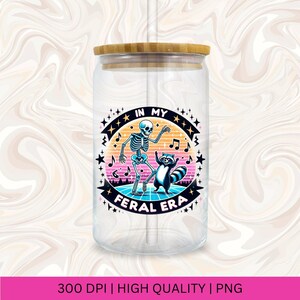 May include: A clear glass tumbler with a bamboo lid and a straw. The tumbler features a colourful graphic of a skeleton and a raccoon dancing with the text "In My Feral Era".