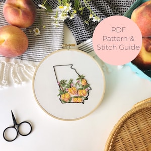 May include: Embroidered hoop art featuring the state of Georgia with peach blossoms and peaches. The hoop is on a white surface with a peach and a white towel in the background. The text "PDF Pattern & Stitch Guide" is on a pink circle.