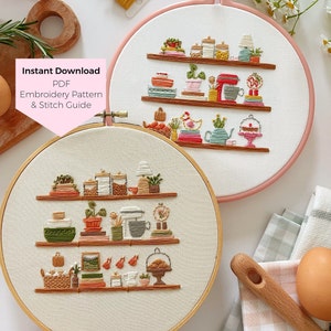 May include: Two embroidery hoop art pieces with a kitchen theme. The hoops are white with a light pink outer ring. The embroidery depicts a kitchen shelf with various items like jars, plants, a mixer, and a cake stand. The text "Instant Download PDF Embroidery Pattern & Stitch Guide" is on a pink background.