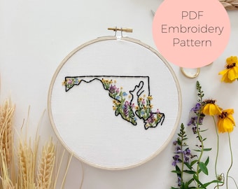 May include: Embroidered floral design of the state of Maryland in a white embroidery hoop. The hoop is on a white surface with dried wheat stalks and flowers.