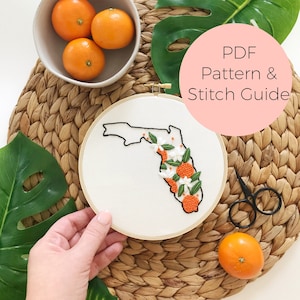 Puede incluir: Arte de bordado en aro con el estado de Florida con flores de azahar y naranjas. El aro está sobre una superficie de paja tejida con una mano sujetando el aro. El texto "PDF Pattern & Stitch Guide" está en un círculo rosa.