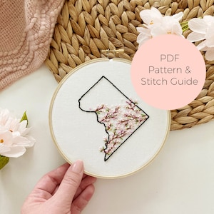 Pode incluir: Arte de bordado em aro que apresenta o contorno do Distrito de Colúmbia com flores de cerejeira rosa e brancas em seu interior. O aro está sobre uma superfície branca com um tapete tecido e uma flor rosa no fundo. O texto "PDF Pattern & Stitch Guide" está em um círculo rosa.