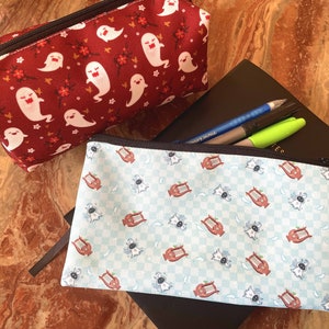 Puede incluir: Dos estuches de lápices con cremallera. Uno es rojo con un patrón de fantasmas blancos y el otro es azul con un patrón de lira blanco y marrón.