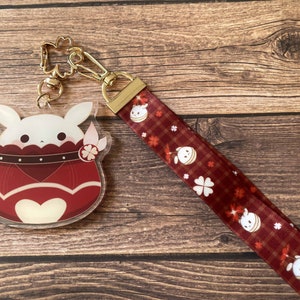 Puede incluir: Un dije acrílico transparente con un personaje de conejo blanco en un diseño de maceta roja, sujeto a un cordón rojo con un patrón repetido del mismo personaje y tréboles. El cordón tiene un cierre y herrajes dorados.