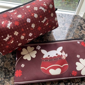 Puede incluir: Dos estuches con cremallera de patrón rojo y blanco. El estuche más pequeño tiene un diseño de trébol blanco y un personaje de dibujos animados blanco con un atuendo rojo y blanco. El estuche más grande tiene un diseño de trébol blanco más pequeño y un personaje de dibujos animados blanco con un atuendo rojo y blanco.