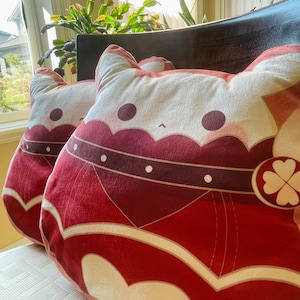 Puede incluir: Dos almohadas de felpa con forma de gato con tela roja, blanca y crema. Las almohadas tienen un diseño de corazón y trébol.
