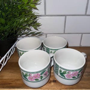 May include: Four white ceramic coffee cups with a floral design featuring pink flowers and green leaves. The cups have a green border and small handles. The cups are on a wooden surface.