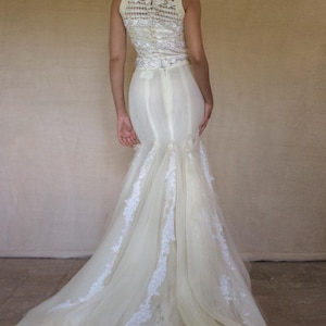 Può includere: Un abito lungo color crema caratterizzato da un corpetto aderente con sovrapposizione di pizzo bianco e chiusura a zip. La gonna si allarga in una silhouette a sirena con pannelli di tulle stratificati e applicazioni in pizzo floreale bianco.