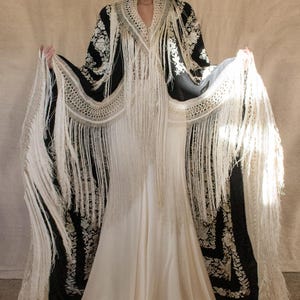 May include: A full-length ivory dress with a black shawl featuring floral embroidery and long, white fringe. The shawl is draped over the shoulders and arms, creating a dramatic silhouette. The dress has a flowing skirt.