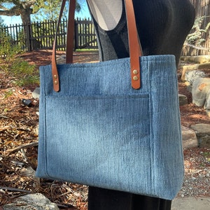 May include: A blue denim tote bag with brown leather straps and gold metal accents. The bag has a large pocket on the front.