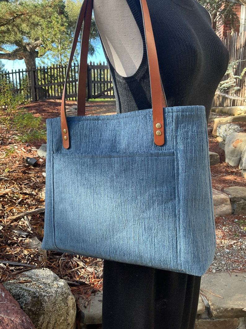 May include: A blue denim tote bag with brown leather straps and gold metal accents. The bag has a large pocket on the front.