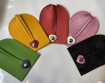 May include: Five leather keychains in different colors, each with a heart-shaped metal charm. The keychains are yellow, red, pink, green, and black.