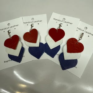 May include: Four leather heart keychains in red, white, and blue. Each keychain has a silver metal ring and a small silver charm. The keychains are displayed on white cards with the text "Handcut Leather" printed on them.
