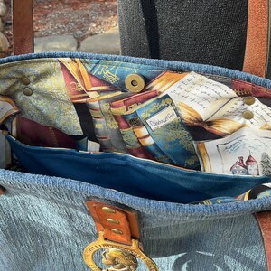 May include: A blue denim tote bag with a brown leather handle and a brass buckle. The inside of the bag is lined with a fabric featuring a pattern of books and a quote that reads "Dunsbonnet Cove".
