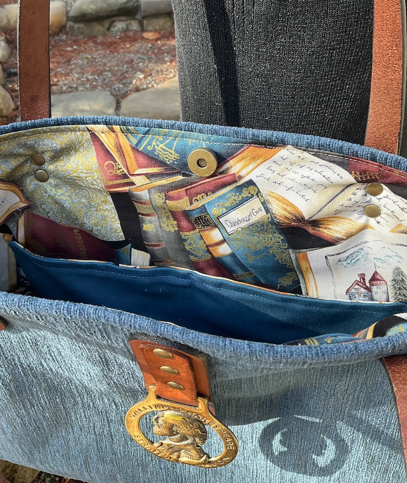 May include: A blue denim tote bag with a brown leather handle and a brass buckle. The inside of the bag is lined with a fabric featuring a pattern of books and a quote that reads "Dunsbonnet Cove".