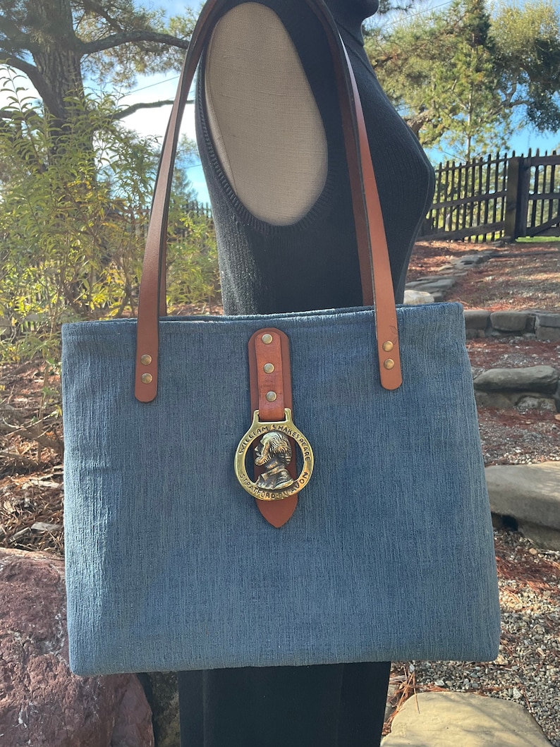 May include: A blue canvas tote bag with brown leather straps and a brass buckle. The buckle has a circular design with a profile of William Shakespeare and the text "William Shakespeare Stratford-upon-Avon".