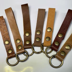 May include: Seven leather keychains with brass rings and two brass rivets. The keychains are in various shades of brown.
