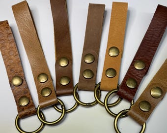 May include: A collection of leather keychains in various shades of brown, tan, and reddish-brown. Each keychain features a leather strap secured with brass-colored rivets and a matching metal ring. The leather textures vary, with some showing embossed patterns.