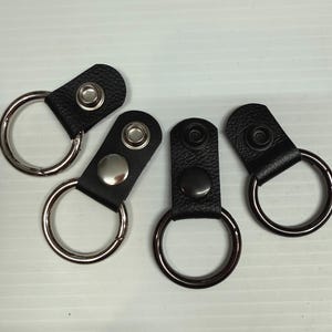 May include: Four black leather keychains with metal snap closures and large silver or black metal rings. The keychains are arranged on a light-colored surface.