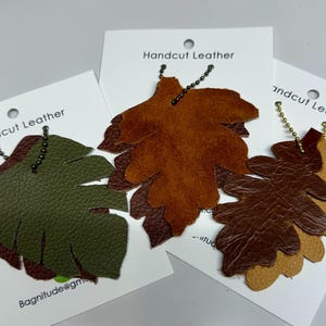 May include: Three leather leaf charms with chains. The leaves are layered and handcut. The leaves are brown, green, and tan. The text "Handcut Leather" is on the white card.
