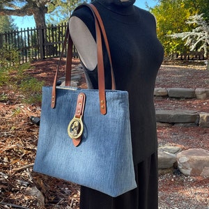 May include: A blue denim tote bag with brown leather straps and a brass buckle. The buckle features a circular design with a raised image of a person's head.