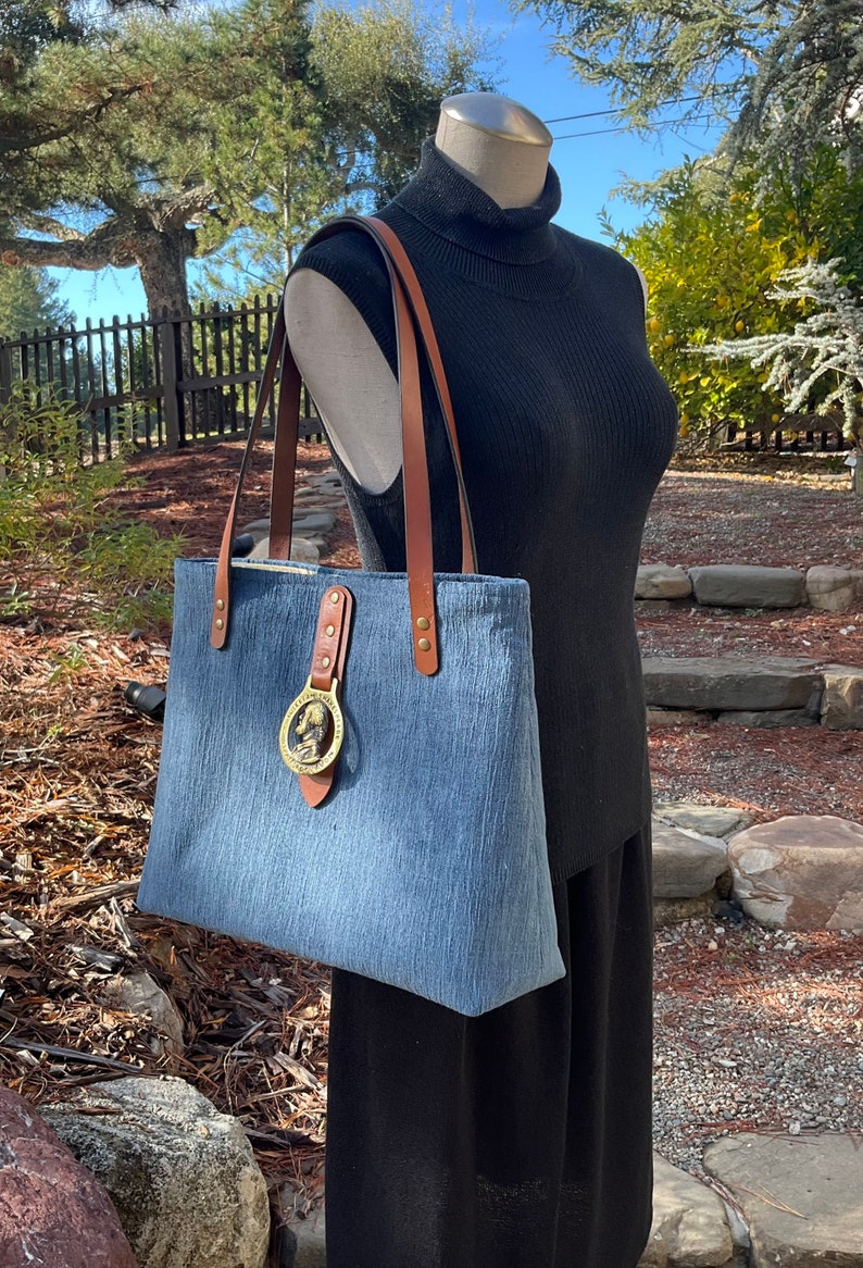 May include: A blue denim tote bag with brown leather straps and a brass buckle. The buckle features a circular design with a raised image of a person's head.