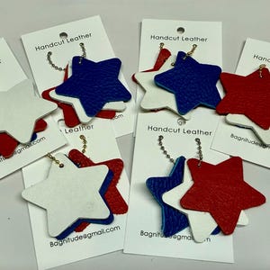 May include: Six leather star keychains in red, white, and blue. Each keychain features three layered stars, with the top star being white, the middle star being blue, and the bottom star being red. The keychains are attached to a gold chain.