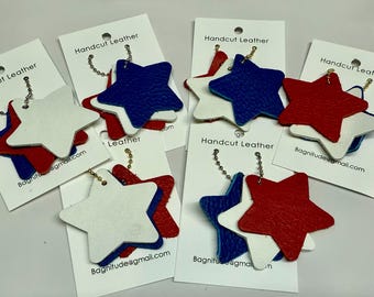 May include: Six leather star keychains in red, white, and blue. Each keychain features three layered stars, with the top star being white, the middle star being blue, and the bottom star being red. The keychains are attached to a gold chain.