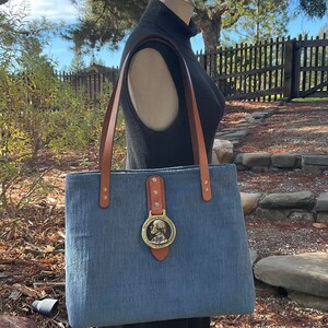 May include: A blue denim tote bag with brown leather straps and a brass buckle featuring a profile of a man. The bag is on a mannequin wearing a black turtleneck.