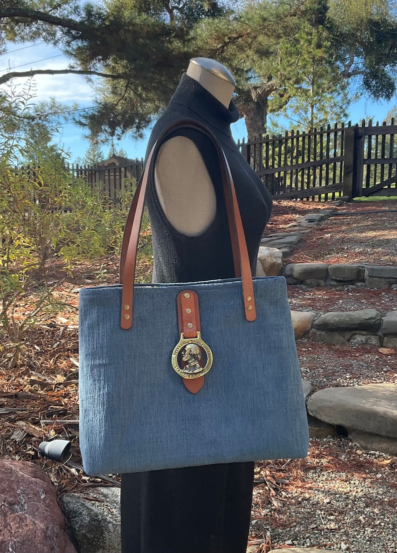 May include: A blue denim tote bag with brown leather straps and a brass buckle featuring a profile of a man. The bag is on a mannequin wearing a black turtleneck.