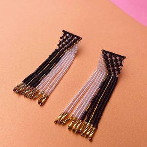 May include: A pair of beaded earrings with a geometric design. The earrings feature black, white, and gold beads. The top portion is a black and white checkered pattern, with long strands of beads hanging down.
