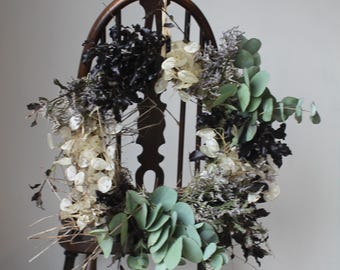 May include: A dried floral wreath with black, white, and green foliage, displayed on a dark wooden chair. The wreath features eucalyptus leaves and various dried flowers, creating a natural, rustic aesthetic.