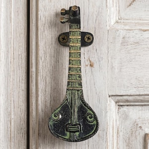 May include: A decorative door knocker shaped like a stringed instrument, featuring a dark green and gold finish. The knocker is mounted on a white wooden door, adding a touch of musical charm to the entrance.