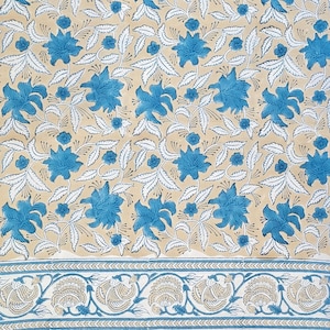 Op de afbeelding: Een textiel met een bloemenpatroon met een herhalend ontwerp van blauwe bloemen en witte bladeren op een beige achtergrond. De onderste rand heeft een gedetailleerd bloemen- en geometrisch patroon in blauw en beige.