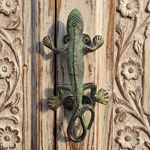 May include: A lizard-shaped metal door handle with a weathered green and bronze finish, featuring a textured scale pattern on its back and four splayed feet. The handle is mounted with visible screws onto a light-colored wooden door panel.