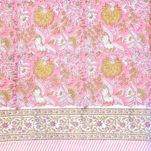 Könnte beinhalten: Rosa Blumengewebe mit einem sich wiederholenden Muster aus großen gelben und weißen Blumen und Blättern. Eine weiße Bordüre mit rosa und goldenen floralen Details und ein rosa-weiß gestreifter unterer Rand.