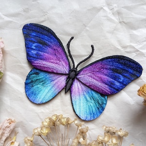 Könnte beinhalten: Ein lila und blauer Schmetterling mit schwarzen Adern und einem schwarzen Körper. Der Schmetterling ist auf einem weißen Stoffhintergrund gestickt.