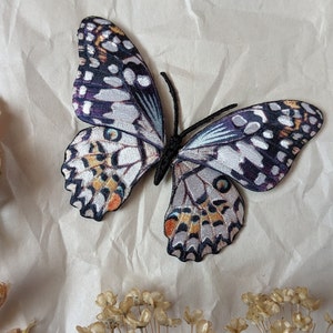 Può includere: Una farfalla bianca e viola con accenti neri e arancioni è ricamata su uno sfondo di tessuto bianco. La farfalla è circondata da fiori secchi.